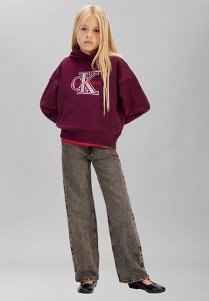 Blondes Kind steht mit den Händen hinter dem Rücken, trägt einen bordeauxfarbenen Calvin Klein Hoodie, graue weite Jeans und schwarze flache Schuhe vor grauem Hintergrund.