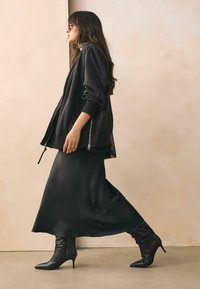 Kvinna iklädd en svart oversized zip-up jacka, svart midiklänning, spetsiga svarta klackstövlar och solglasögon som går inomhus.