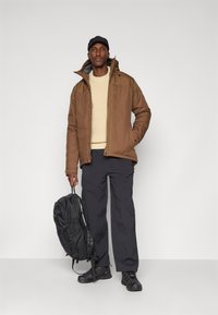 Brun isoleret jakke, beige strikket sweater, mørke bukser, sort rygsække og sorte sko. Jakken har en høj krave og synlig syning.