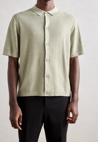 Korte mouw, lichtgroen gebreid shirt met een kraag, voorzien van een knoopsluiting aan de voorkant en een geribbelde textuur, gecombineerd met zwarte broek.