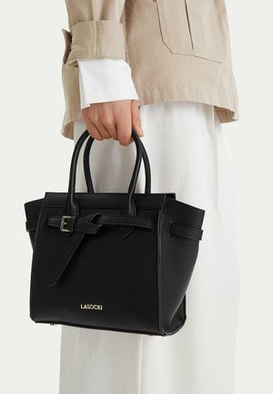 Persona sosteniendo un bolso de cuero negro Lasocki con doble asa, vistiendo una chaqueta beige y pantalones blancos.