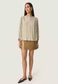 Blusa beige a maniche lunghe con un motivo texturizzato; abbinata a una mini gonna in pelle marrone e a delle ballerine nere a punta. Design semplice, vestibilità rilassata.