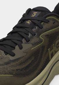 Κοντινό πλάνο ενός μαύρου και ελαιοπράσινου αθλητικού παπουτσιού με λεπτομερή ύφασμα από δίχτυ, μαύρα κορδόνια και λογότυπο στη γλώσσα και την πλευρά.