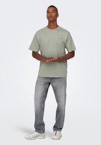 Camiseta de algodón gris claro, corte relajado, con mangas cortas y un sutil logotipo en azul. Combinada con jeans gris claro y zapatillas blancas.