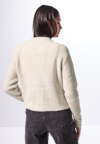 Beige ribbad tröja med hög hals och långa ärmar, kombinerad med mörkgrå byxor. Betonar textur och strukturerad passform.