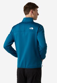 Giacca soft shell color teal con colletto alto, tasche con zip e un piccolo logo bianco sulla parte superiore della schiena. Leggera, con una superficie liscia.