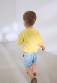 Bambino con maglietta gialla a maniche corte e pantaloncini a quadretti blu, che cammina su un pavimento chiaro con ombre morbide. Sandali marroni ai piedi.
