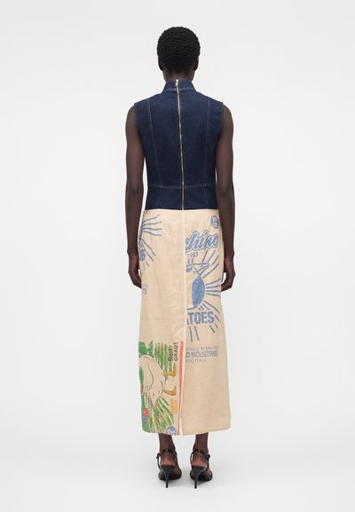 Λεπτό άτομο που φοράει αμάνικο φόρεμα με σκούρο τζιν πάνω μέρος και μπεζ φούστα με πολύχρωμο γραφικό κείμενο και εικόνες, στέκεται με την πλάτη στην κάμερα.