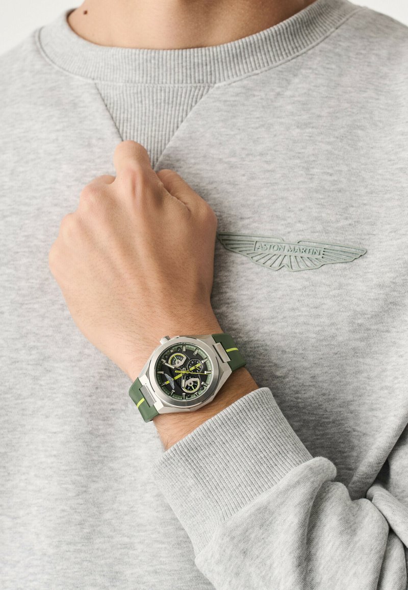 Primer plano de una muñeca que lleva un reloj cronógrafo plateado con una correa verde y amarilla, apoyada sobre una sudadera gris de Aston Martin.