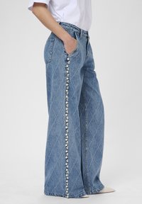 Široké modré džínsy s diamantovým prešívaním a dekoratívnym perlovým a korálkovým lemom pozdĺž vonkajších švov, nosené s bielou košeľou a béžovými topánkami.