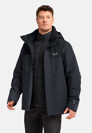 Uomo che indossa giacche invernali nere Jack Wolfskin a strati, guardando leggermente verso l’alto e di lato, in piedi davanti a uno sfondo chiaro e semplice.