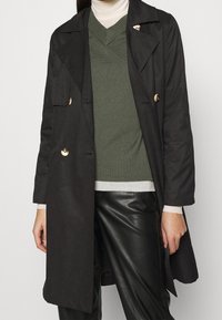 Černý trench coat se zlatými knoflíky, dlouhými rukávy a bočními kapsami, nošený přes olivově zelený svetr a černé kožené kalhoty.