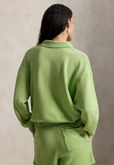 Kvinde med mørkt hår iført lysegrøn langærmet skjorte og matchende shorts med baglomme, stående med hænderne i lommerne og vendt væk.