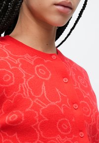 Rode gebreide cardigan met bloemenmotieven, een ronde hals en vijf knopen aan de voorkant. De stof lijkt zacht te zijn met een gestructureerde afwerking.