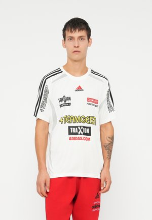 Maglietta bianca a maniche corte con strisce nere Adidas, con loghi colorati in nero, giallo e rosso. Indossata con pantaloni rossi.