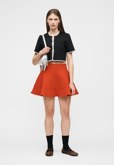 Γυναίκα που φοράει μαύρο κοντό πουλόβερ με άσπρη διακόσμηση, κόκκινη φούστα με καμπάνα, μαύρες κάλτσες, παπούτσια με σχεδιασμό λεοπάρδαλης και κρατά μια ασημένια τσάντα με φουντίτσες.