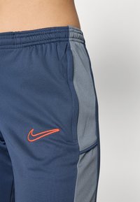 Navy treningsbukser med grå mesh-sidelapper. Har en oransje Nike-logo på låret. Elastisk midjebånd for justerbar passform.