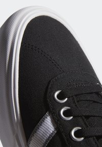 Musta kangaskenkä valkoisella kumipohjalla, jossa on hopeiset silmukat, mustat nauhat ja sivuvalkoinen raita. Lähikuva korostaa tekstuuria.