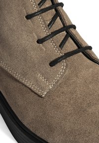 Braune Wildleder-Ankle-Boots mit schwarzen Schnürsenkeln, kontrastierender beiger Naht und strukturierter Oberfläche. Gummisohle mit leichtem Höhenunterschied.
