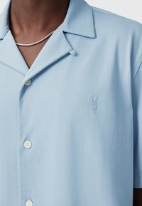 Camicia azzurra a maniche corte realizzata in tessuto morbido, con colletto a punta, bottoni bianchi e un piccolo logo ricamato sul petto.
