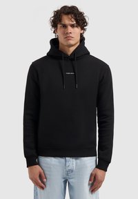Zwarte hoodie van zachte stof, met een voorzak, trekkoord en kleine witte tekst op de borst. Gecombineerd met lichtblauwe jeans.