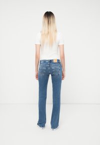 Sieviete ar gariem blondiem ombre matiem, tērpusies baltā īsām piedurknēm topā, zilo džinsu biksēs un baltās sporta apavos, stāv ar muguru pret vienkāršu balto fona.