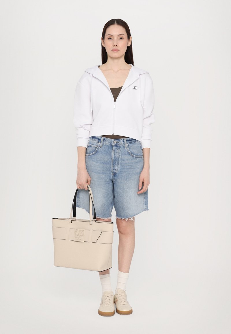 Felpa bianca tagliata, canotta oliva, pantaloni corti in denim strappati, borsa tote beige con accenti neri, sneakers beige, calzini bianchi, modello in piedi.