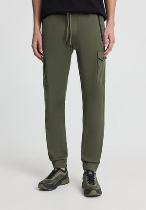 Uomo che indossa pantaloni cargo jogger verde oliva con vita con coulisse e polsini elasticizzati, abbinati a sneaker coordinate verde oliva e nere.