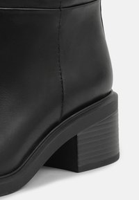 Gros plan sur l'arrière et le talon d'une botte cheville en cuir noir avec un talon bloc empilé et une finition lisse.