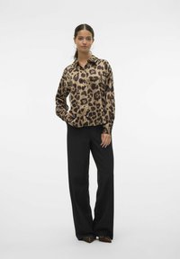 Blusa a stampa leopardata in beige e marrone, con colletto e bottoni, abbinata a pantaloni larghi neri a vita alta e scarpe marroni a punta.