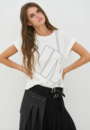 Hvid T-shirt med korte ærmer og sort kantet grafisk design; båret med sort plisseret nederdel med bredt bælte og spænde detaljer.