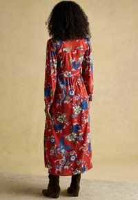 Rotes Blumen-Kleid mit langen Ärmeln, das blaue und cremefarbene Blumenmuster zeigt, mit einer taillierten Taille und einem fließenden Rock, kombiniert mit braunen Absatzstiefeln.