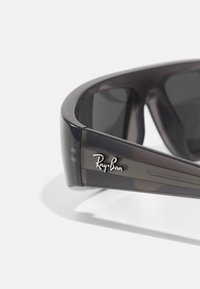 Sončna očala Ray-Ban s polprosojnim temno sivo okvirjem in temnimi lečami. Značilnosti vključujejo kovinski logotip na roki in eleganten, oglat oblikovan dizajn.