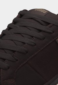 Κοντινό πλάνο σκούρου καφέ αθλητικού παπουτσιού με χοντρές κορδόνια, σουέτ υφή και ραμμένες λεπτομέρειες στην επάνω πλευρά.