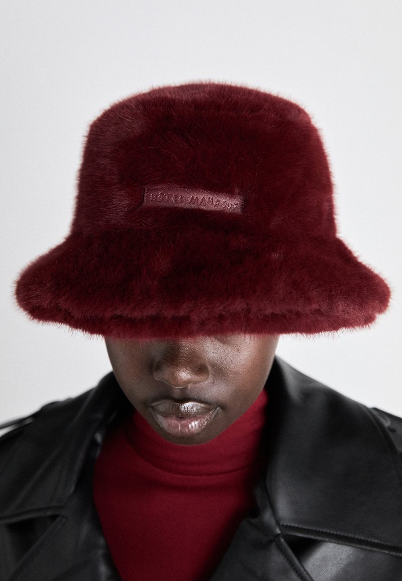 Burgunderfarget faux fur bøttehatt med bred brem og lær merkeetikett. Myk tekstur og rund form som dekker øynene.