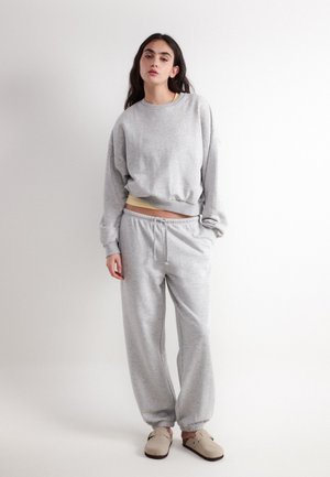 Jeune femme portant un sweat-shirt gris clair et un pantalon de survêtement assorti avec des chaussures beige à enfiler, debout devant un fond blanc.