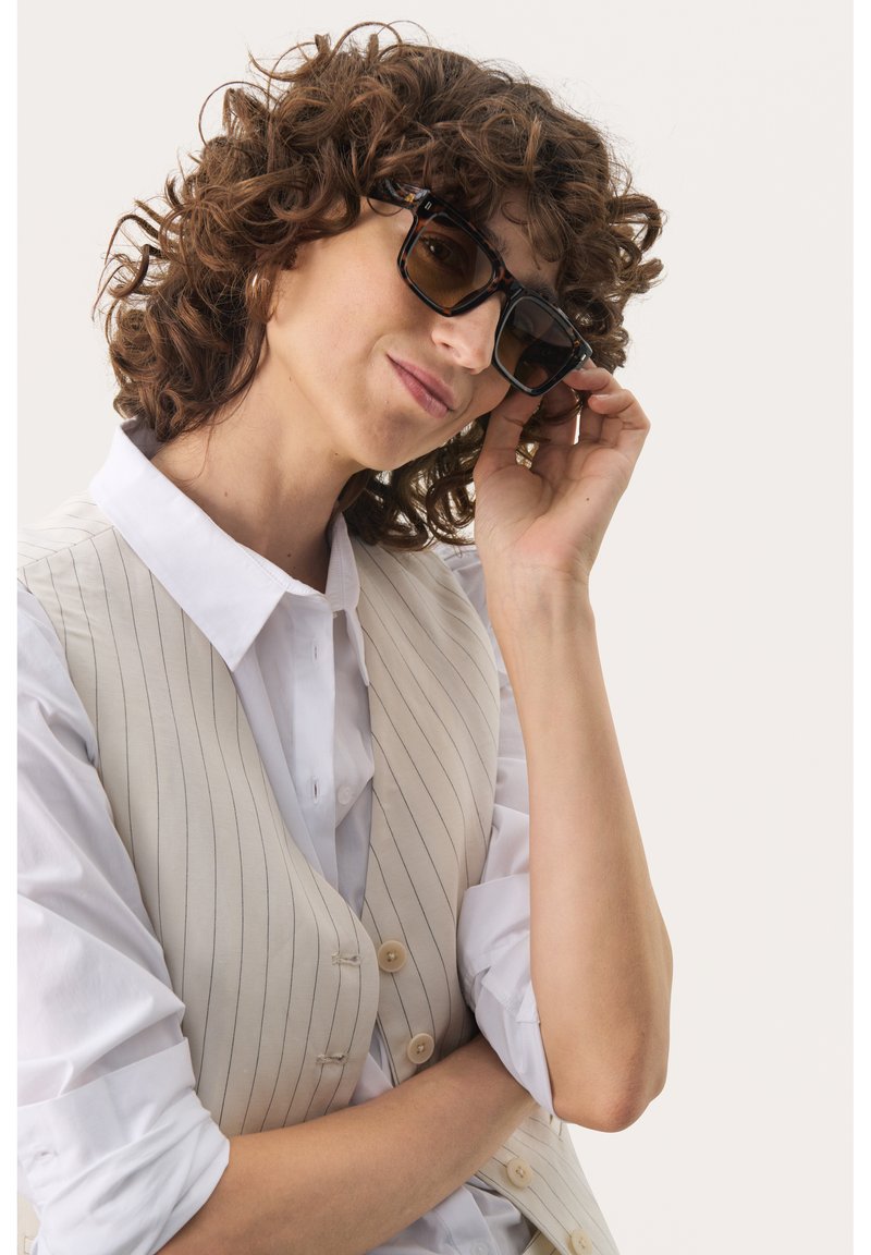 Occhiali da sole con una montatura spessa in tartaruga nera e marrone, dotati di lenti scure. Indossati con una camicia bianca e un gilet crema.