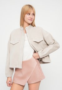 Veste en velours côtelé beige avec deux poches poitrine et un col côtelé, associée à une jupe portefeuille rose clair en tissu doux et texturé.