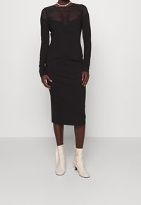 Robe midi noire à manches longues avec panneau en maille transparente au niveau de l'encolure, silhouette ajustée, assortie à des bottines crème.