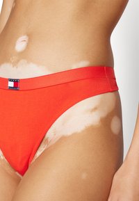 Rode katoenen bikini onderkant met een gladde textuur en een gebrandmerkte tailleband. Zichtbaar huidpigmentatiecontrast aan de zijkant.