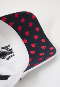 Gorra blanca con un logotipo bordado en negro. La parte inferior del visor es de color marino con un patrón repetido de manzanas rojas. Textura de tela suave.