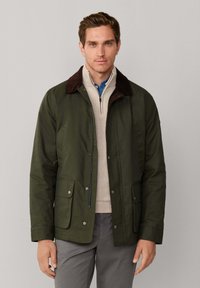 Chaqueta verde oscuro con cuello de pana marrón, cremallera frontal y botones a presión. Cuenta con dos bolsillos frontales con solapas planas. Acabado de tela texturizada.