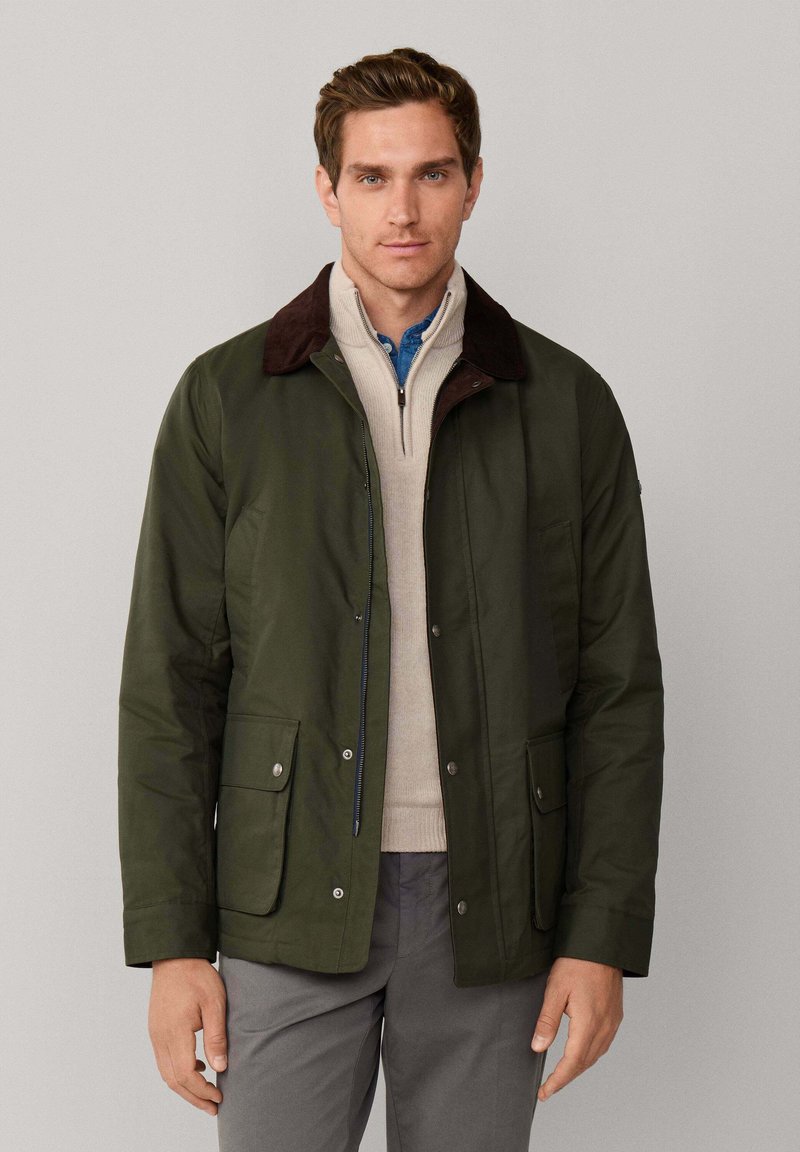 Chaqueta verde oscuro con cuello de pana marrón, cremallera frontal y botones a presión. Cuenta con dos bolsillos frontales con solapas planas. Acabado de tela texturizada.
