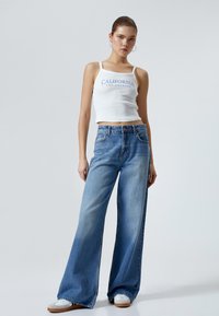 Lysblå vide jeans med høj talje, lavet af denim. Bæres med en hvid kort tanktop med blå tekst. Hvite sneakers.