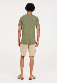 Olivinės spalvos marškinėliai su mažomis, spalvingomis valčių ir medžių pynėmis, derinami su šviesiai rudos spalvos kelnių šortais su šoniniais kišeniais. Rodyklė iš nugaros.