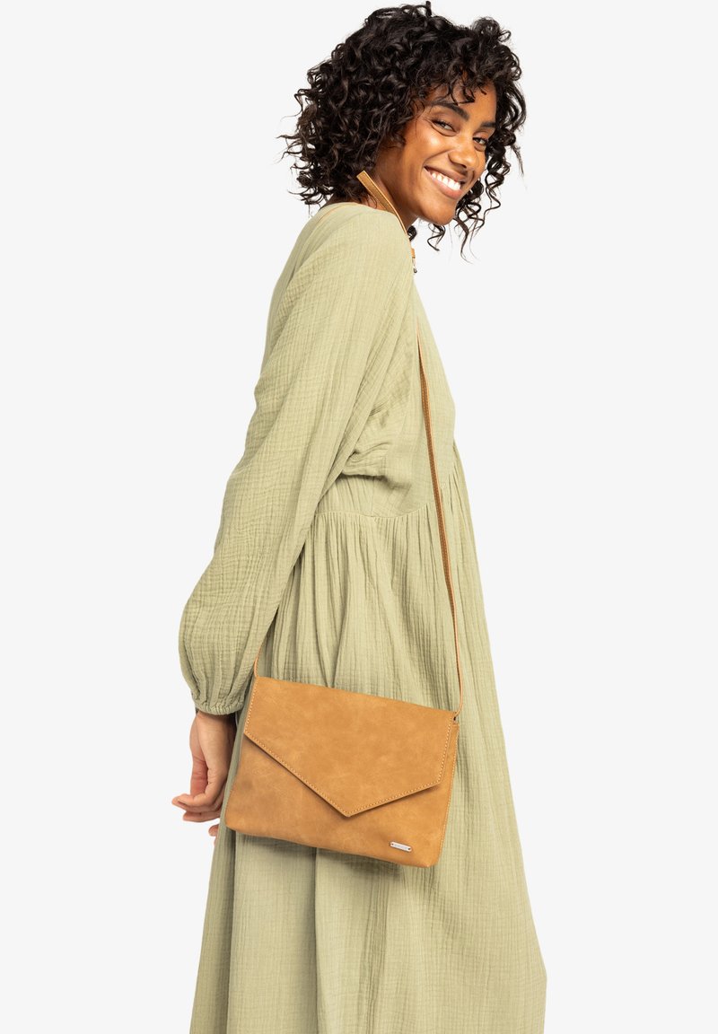 Femme souriante avec des cheveux bouclés, portant une robe longue à manches longues vert clair et tenant un sac bandoulière beige avec une fermeture à rabat.