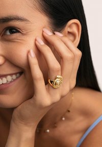 Anello d'oro con un design a rosa, indossato sulla mano. Texture liscia con dettagli intricati. Sfondo sfocato con illuminazione morbida.