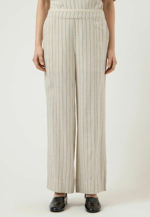 Kvinde iført beige bukser med brede ben og tynde lodrette sorte striber samt sorte laksko, stående mod en neutral baggrund.