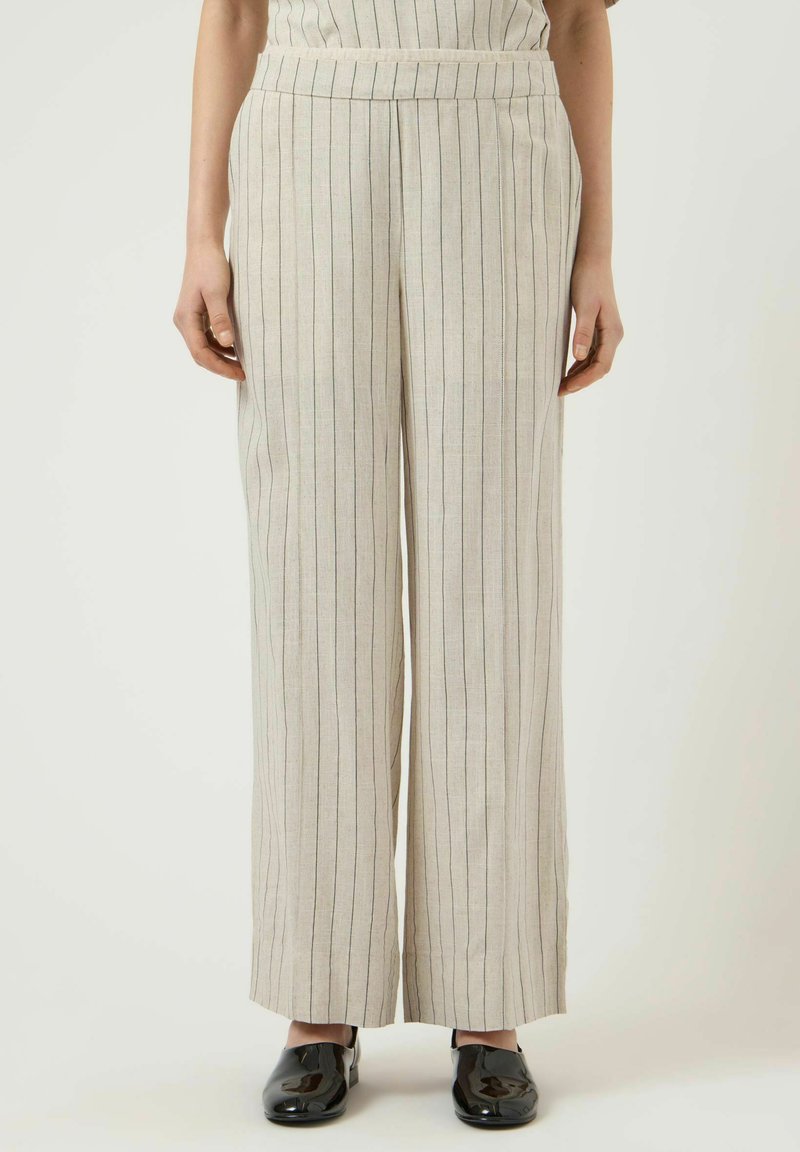 Femme portant un pantalon beige à jambes larges avec de fines rayures verticales noires et des chaussures en cuir verni noir, debout devant un fond neutre.