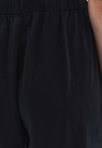 Svart, lätt tyg med en slät textur, som har ett elastiskt midjeband och avslappnad passform. Delvis vy av byxor från midjan och ner.